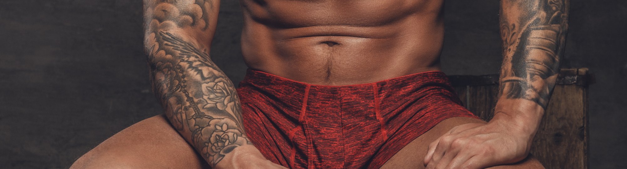 muskulöser, tätowierter Mann in roten Shorts sitzt vor dunklem Hintergrund