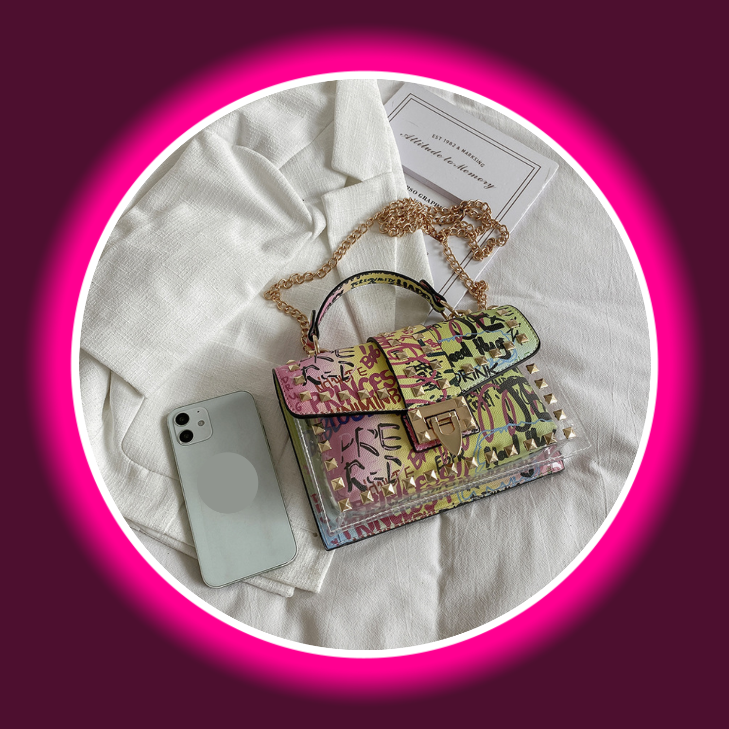bunte Handtasche mit Graffiti-Druck, Tragegriff und goldfarbenen Details, auf weißem Tuch liegend, daneben ein Smartphone, Sexymize